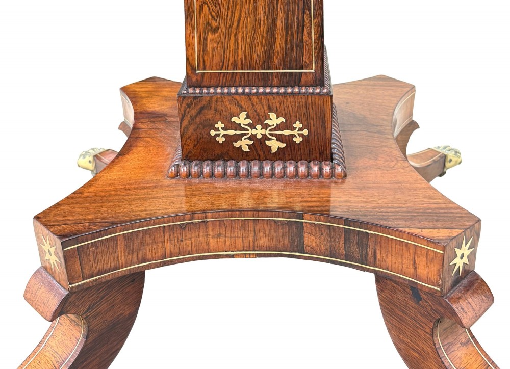 A circular Regency rosewood centre table featuring brass inlay motifs around the rim, a central pedestal support, and a quadform base with splayed legs ending in brass paw castors. View X