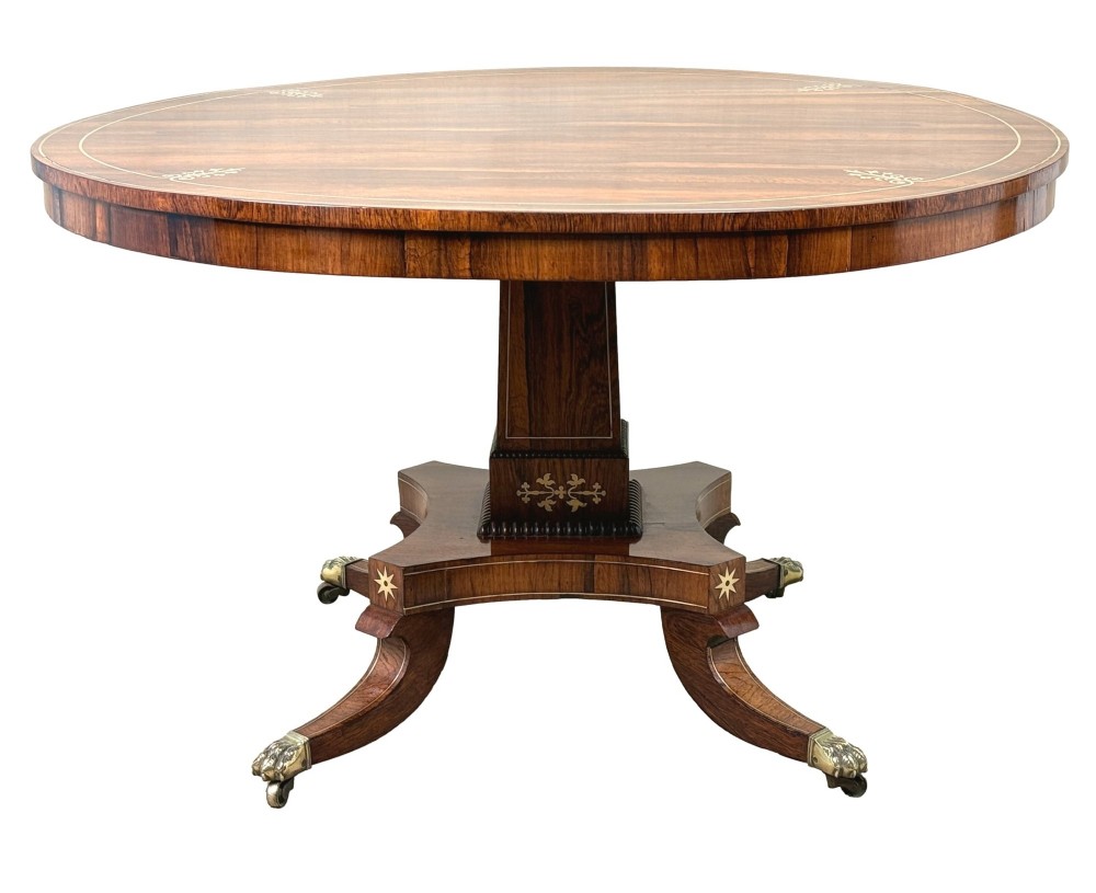 A circular Regency rosewood centre table featuring brass inlay motifs around the rim, a central pedestal support, and a quadform base with splayed legs ending in brass paw castors. View XIII