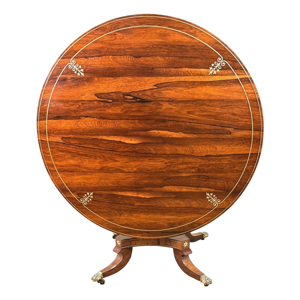 A circular Regency rosewood centre table featuring brass inlay motifs around the rim, a central pedestal support, and a quadform base with splayed legs ending in brass paw castors. View XIV
