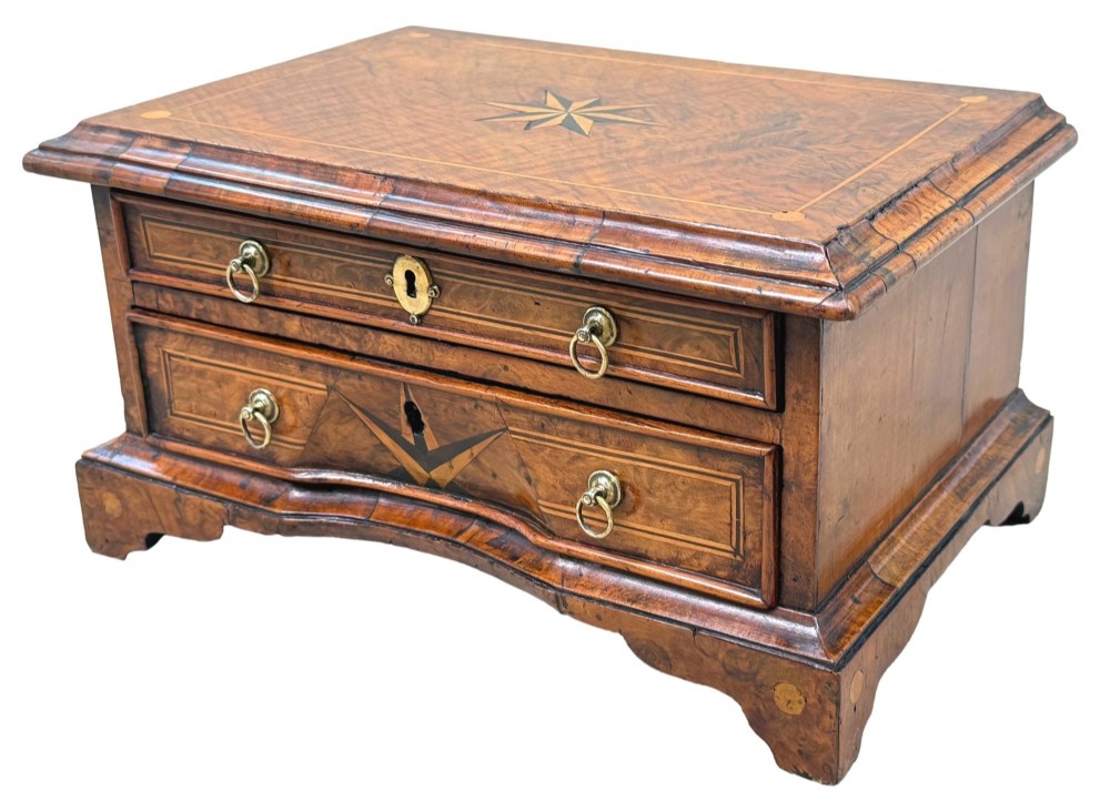 An early 18th-century walnut miniature chest featuring two drawers, star marquetry inlay, crossbanding, brass drop ring handles, and bracket feet.