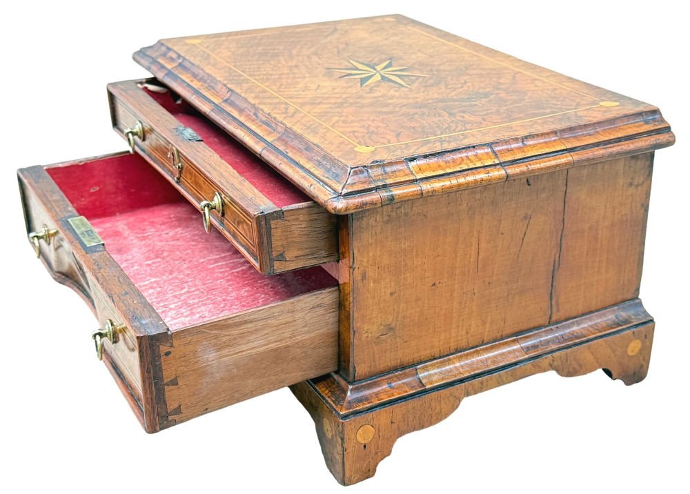 An early 18th-century walnut miniature chest featuring two drawers, star marquetry inlay, crossbanding, brass drop ring handles, and bracket feet. View II