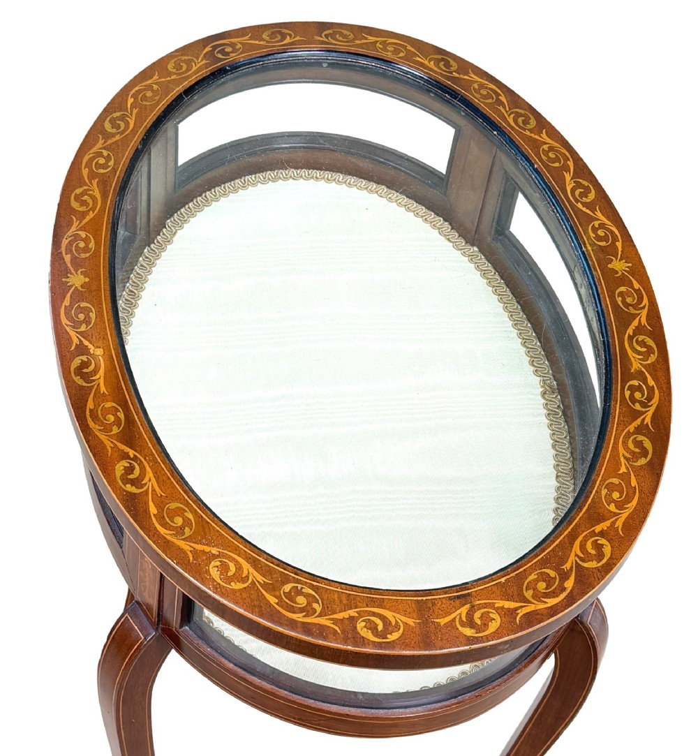 An oval late 19th-century mahogany bijouterie display table featuring glass side panels, inlaid floral marquetry, slender cabriole legs, and a cross stretcher. View IX
