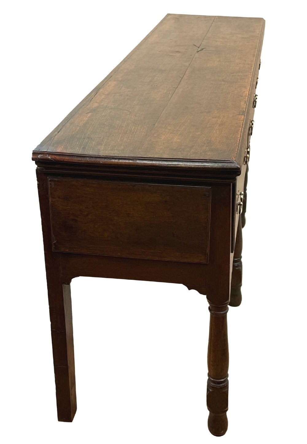 An early eighteenth-century oak dresser base featuring three drawers with brass drop handles, brass keyhole escutcheons, turned front legs, and a dark brown finish. View VI