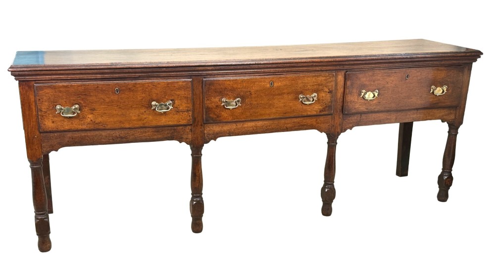 An early eighteenth-century oak dresser base featuring three drawers with brass drop handles, brass keyhole escutcheons, turned front legs, and a dark brown finish. View VIII