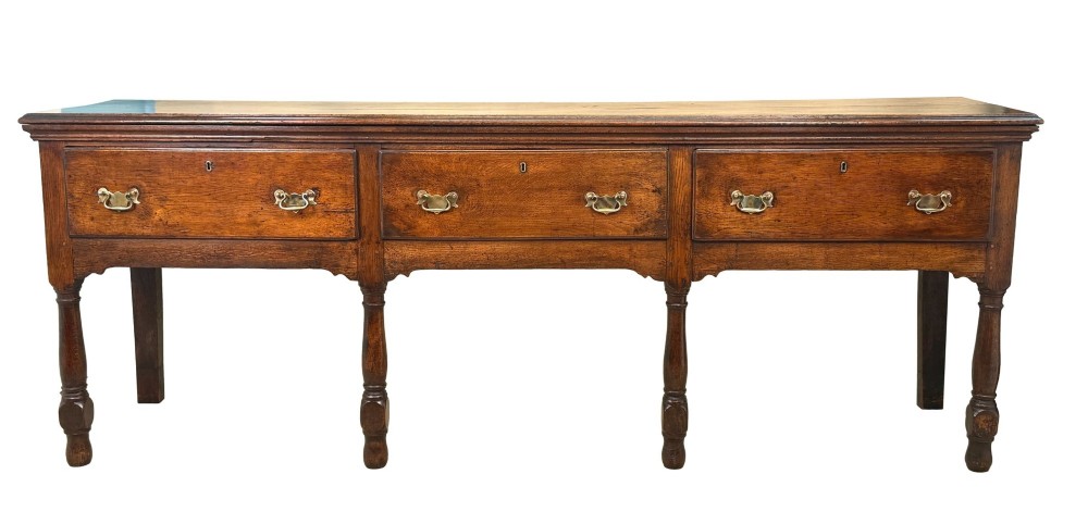 An early eighteenth-century oak dresser base featuring three drawers with brass drop handles, brass keyhole escutcheons, turned front legs, and a dark brown finish. View X