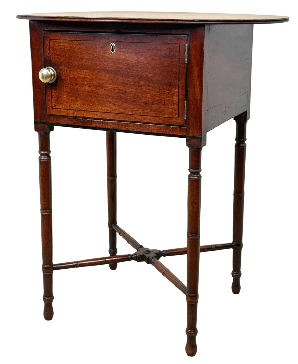 An early 19th-century Georgian mahogany side table featuring an oval top, single door cupboard with brass knob, turned legs, and an X-shaped cross stretcher.