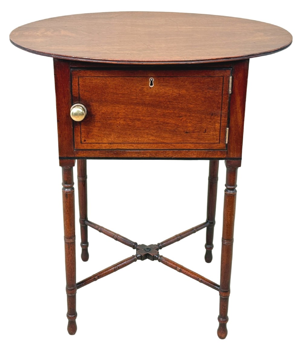 An early 19th-century Georgian mahogany side table featuring an oval top, single door cupboard with brass knob, turned legs, and an X-shaped cross stretcher. View X
