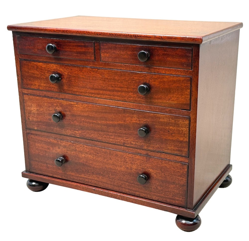A mid-nineteenth-century miniature mahogany chest of drawers featuring two short over three long drawers, turned dark knobs, and bun feet.
