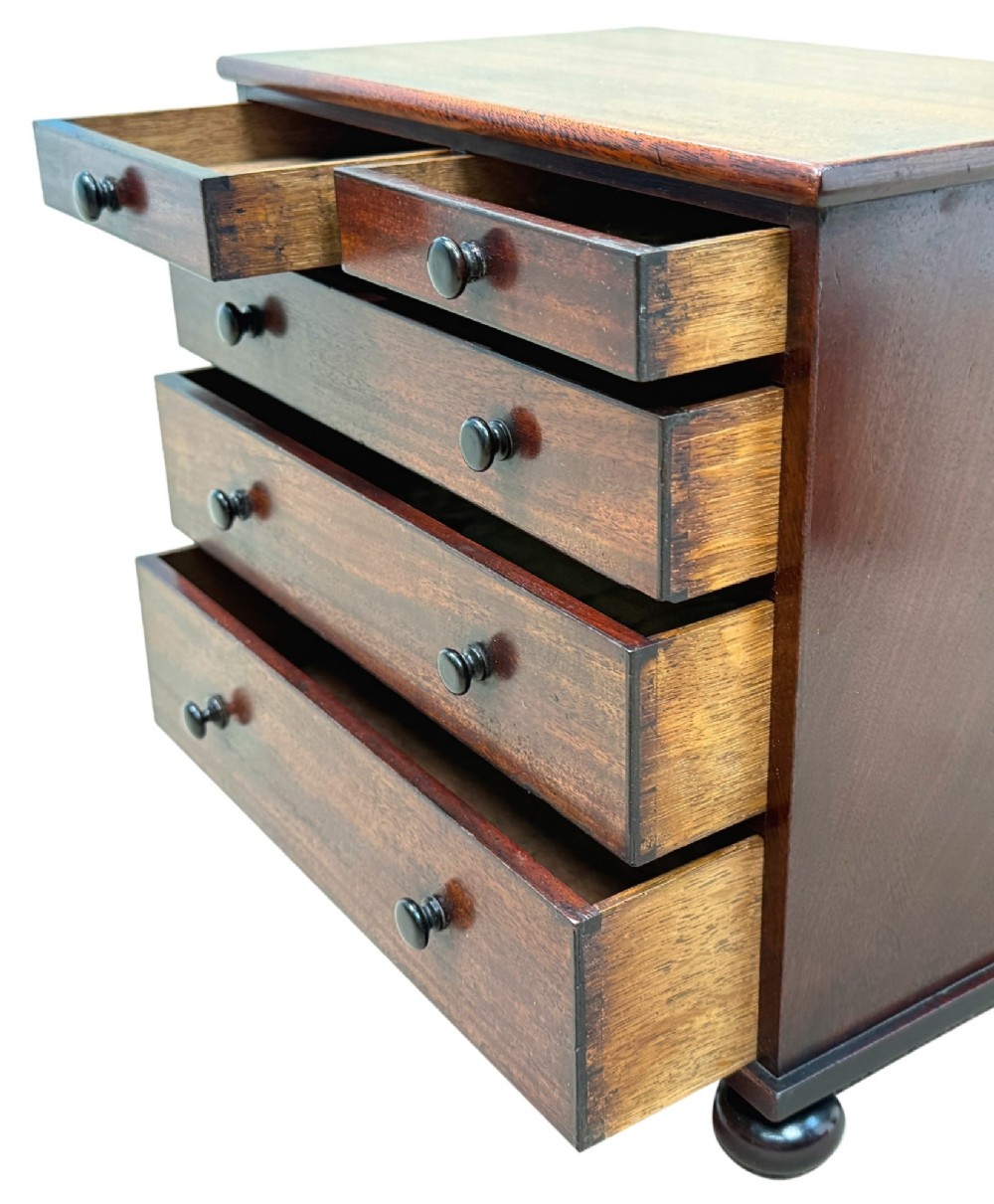 A mid-nineteenth-century miniature mahogany chest of drawers featuring two short over three long drawers, turned dark knobs, and bun feet. View VI