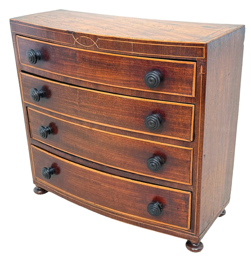 An 18th-century Georgian mahogany miniature bowfront chest of drawers featuring boxwood inlay, four graduated drawers with turned wooden knobs, and low bun feet.