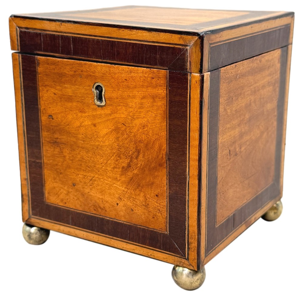 A square Georgian satinwood tea caddy featuring dark wood crossbanded borders, a central brass keyhole escutcheon, and four brass ball feet.