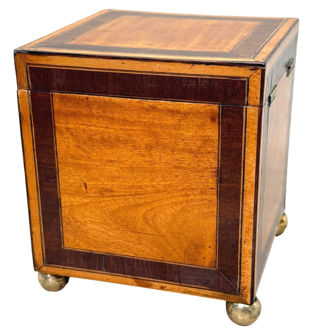 A square Georgian satinwood tea caddy featuring dark wood crossbanded borders, a central brass keyhole escutcheon, and four brass ball feet. View II