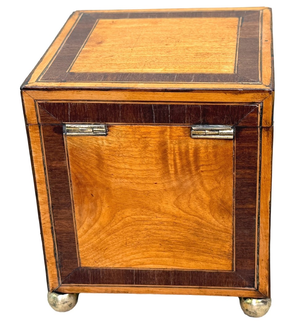 A square Georgian satinwood tea caddy featuring dark wood crossbanded borders, a central brass keyhole escutcheon, and four brass ball feet. View III