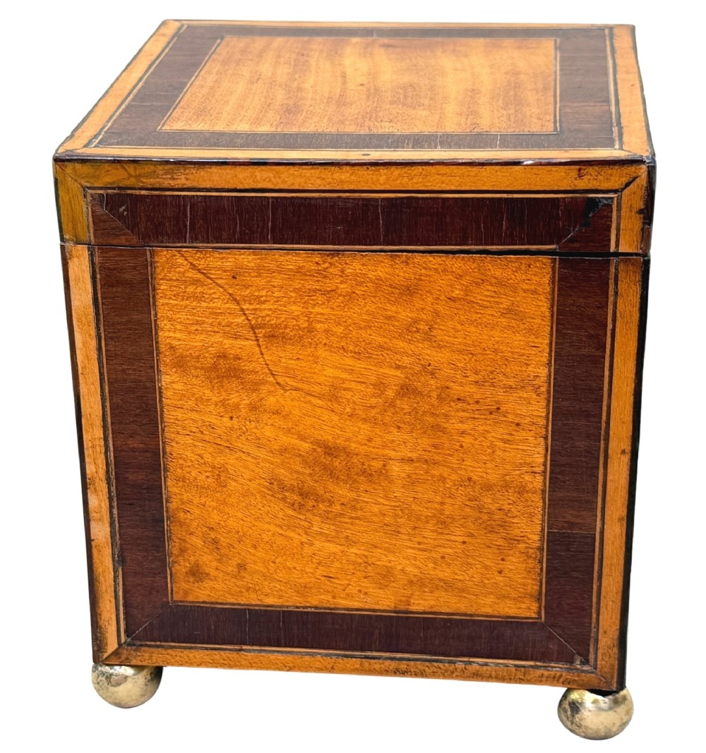 A square Georgian satinwood tea caddy featuring dark wood crossbanded borders, a central brass keyhole escutcheon, and four brass ball feet. View V