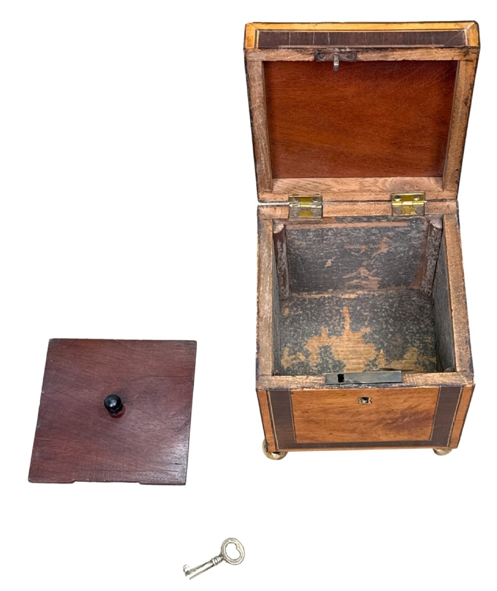 A square Georgian satinwood tea caddy featuring dark wood crossbanded borders, a central brass keyhole escutcheon, and four brass ball feet. View VI