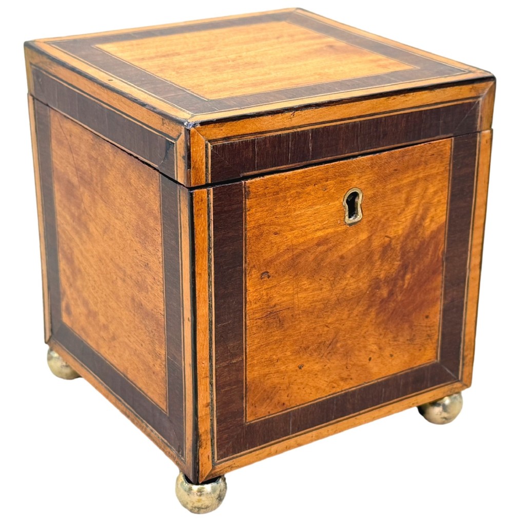 A square Georgian satinwood tea caddy featuring dark wood crossbanded borders, a central brass keyhole escutcheon, and four brass ball feet. View VII