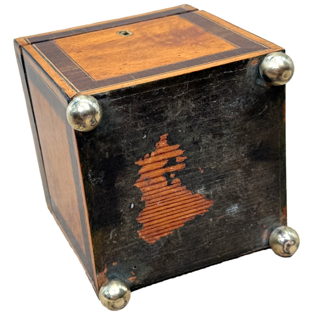 A square Georgian satinwood tea caddy featuring dark wood crossbanded borders, a central brass keyhole escutcheon, and four brass ball feet. View VIII