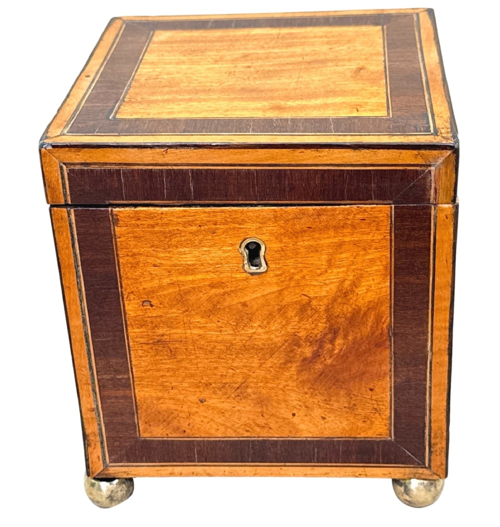 A square Georgian satinwood tea caddy featuring dark wood crossbanded borders, a central brass keyhole escutcheon, and four brass ball feet. View IX