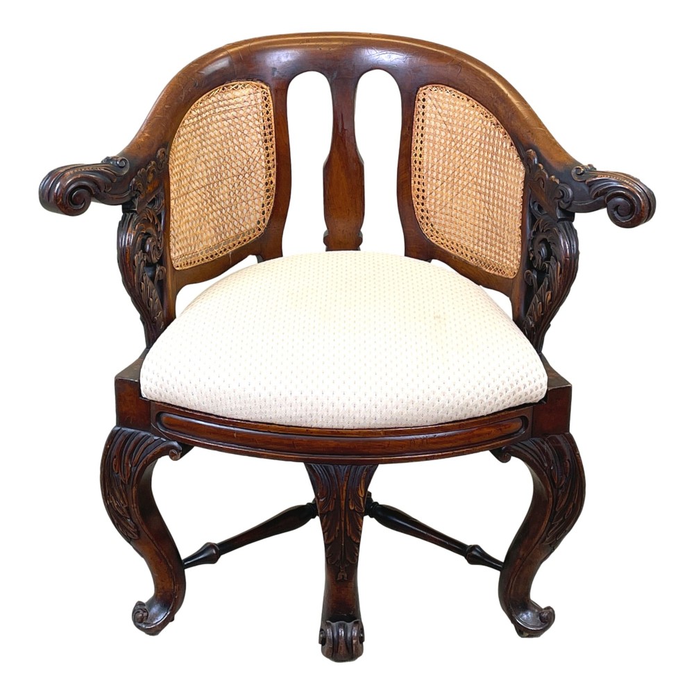 A 19th-century mahogany Burgermeister desk chair featuring a curved backrest with woven cane panels, carved armrests, an upholstered cream seat, and cabriole legs. View VI