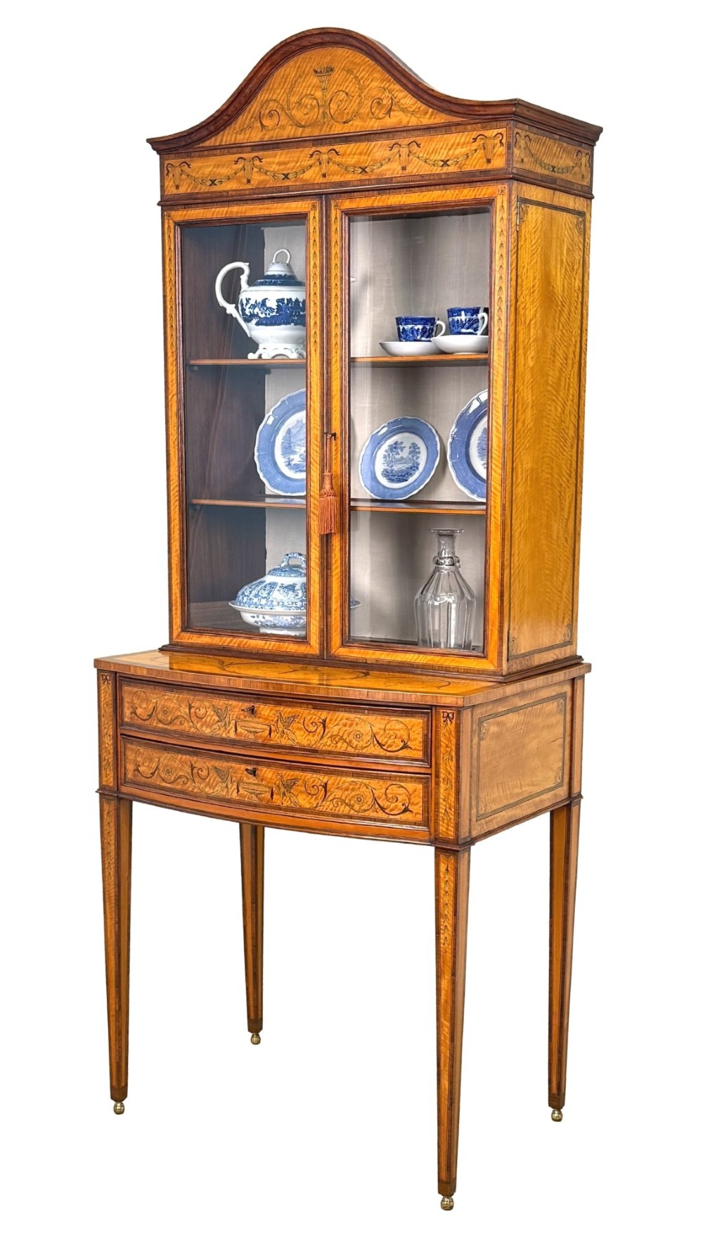 A 19th-century inlaid satinwood display cabinet featuring an arched cresting, glazed upper doors, marquetry frieze drawers, and square tapered legs.