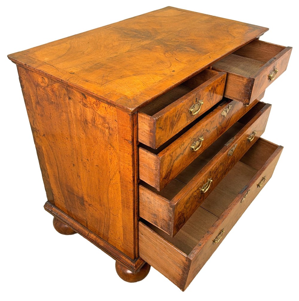 An early eighteenth-century Queen Anne walnut chest of drawers featuring two short over three long graduated drawers, brass handles, pierced escutcheons, and turned bun feet. View V