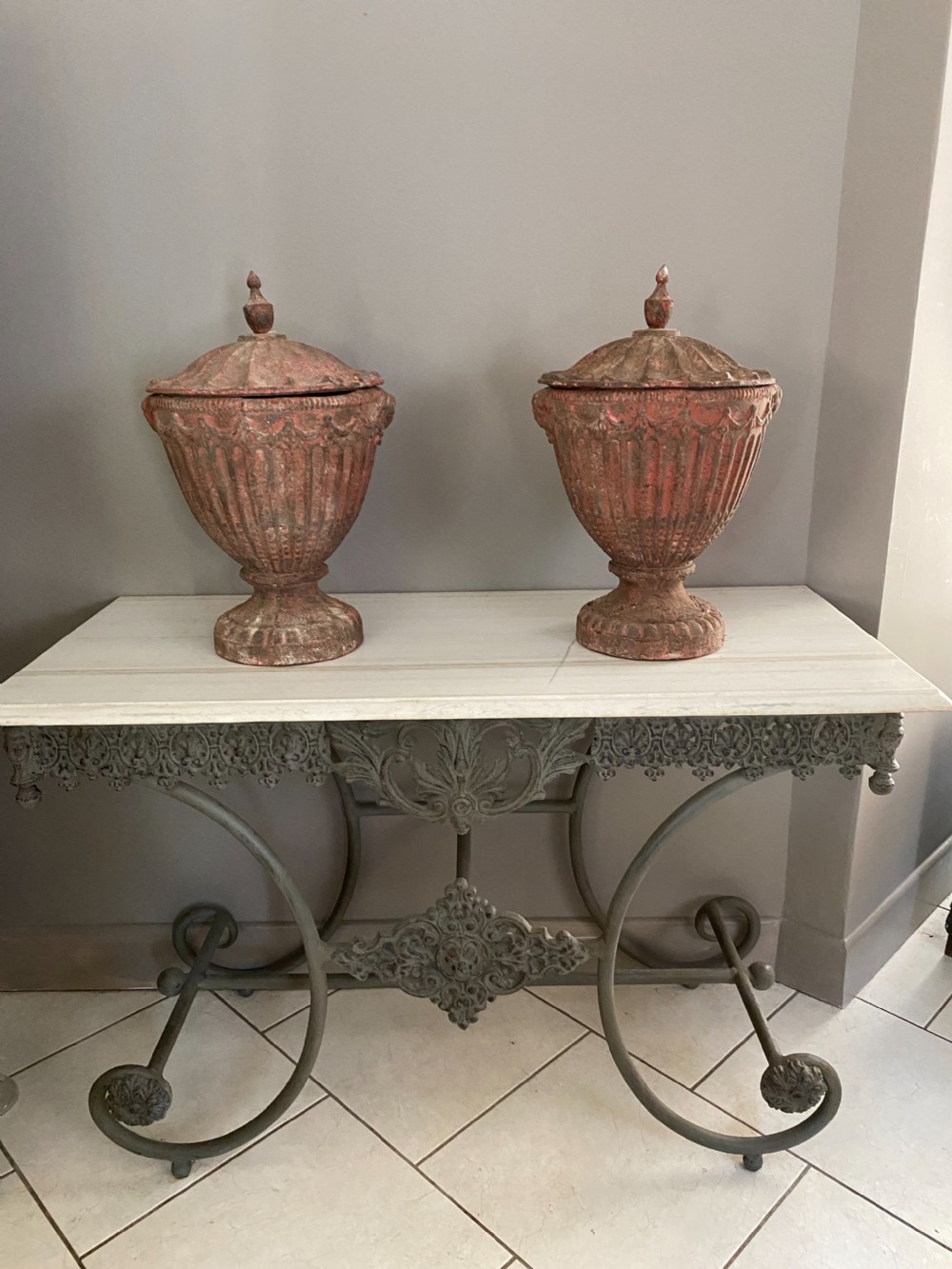 A pair of antique cast iron urns with fluted bodies, finial-topped lids, and weathered red patinated surfaces resting on pedestal bases.