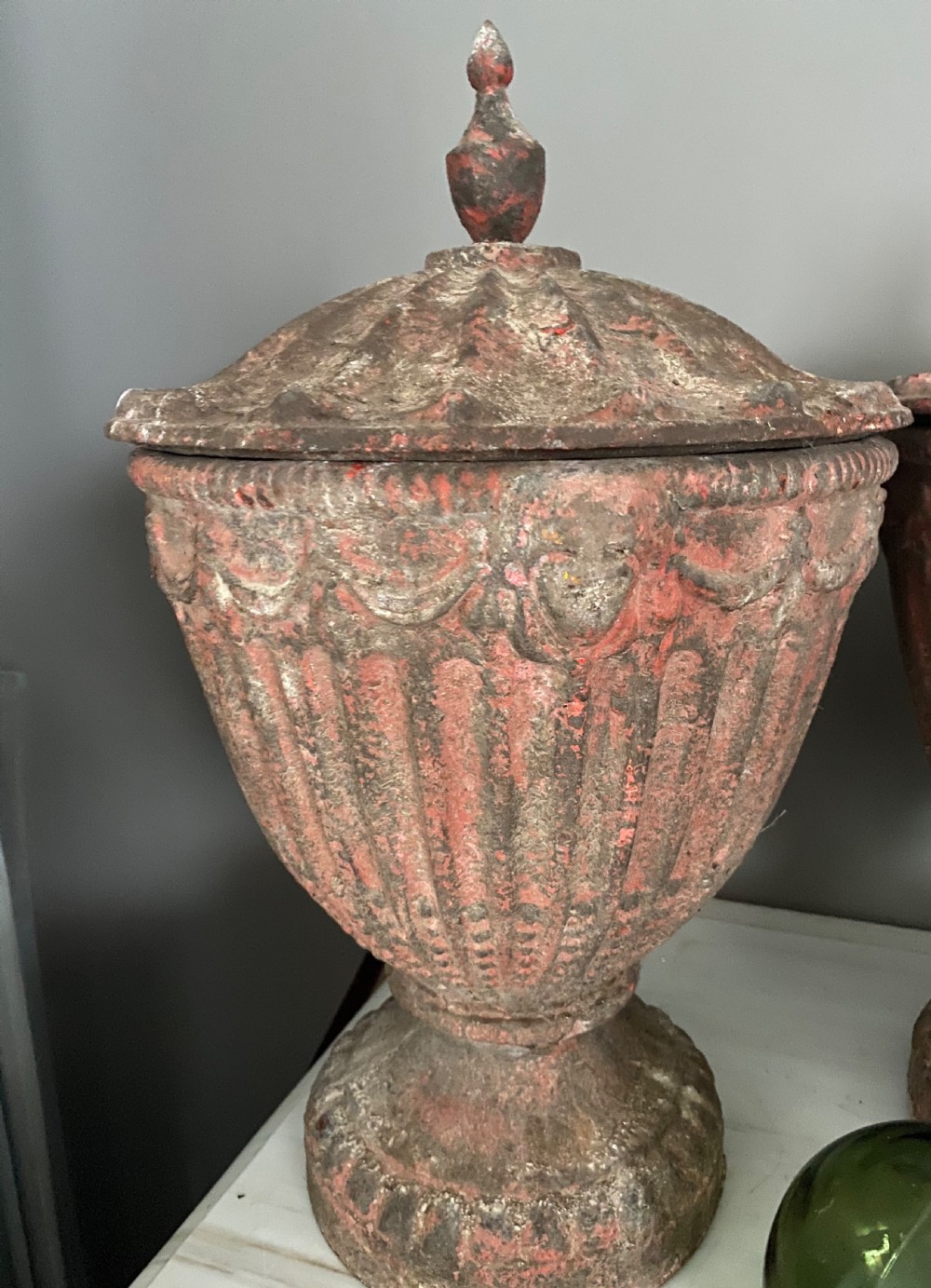 A 19th-century fluted cast iron urn featuring weathered terracotta paint, draped festoon motifs, and a pointed lid on a pedestal base.