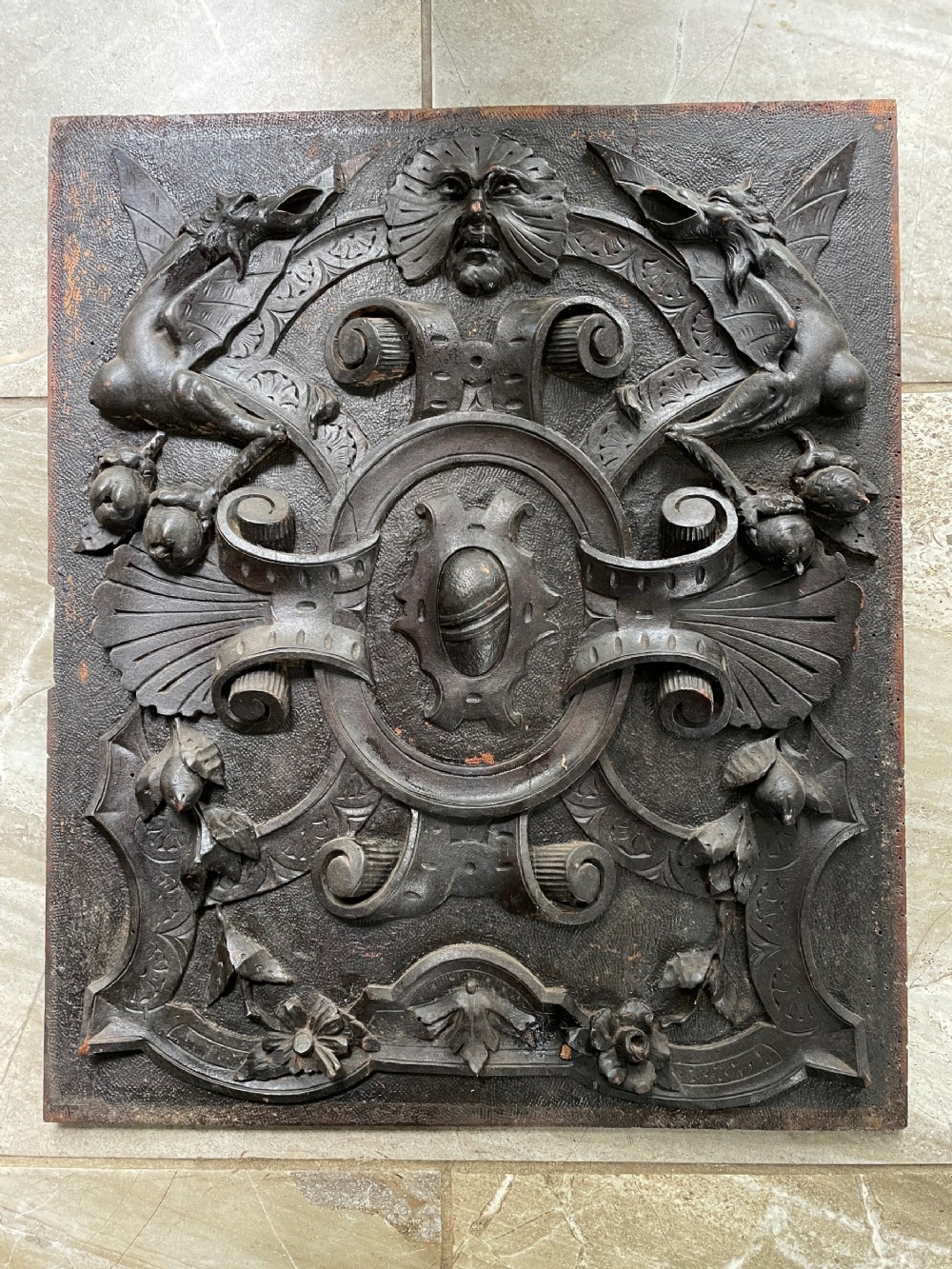 A rectangular dark walnut wood panel featuring high-relief carvings of a Green Man mask, mythical winged beasts, scrolls, and foliate motifs.