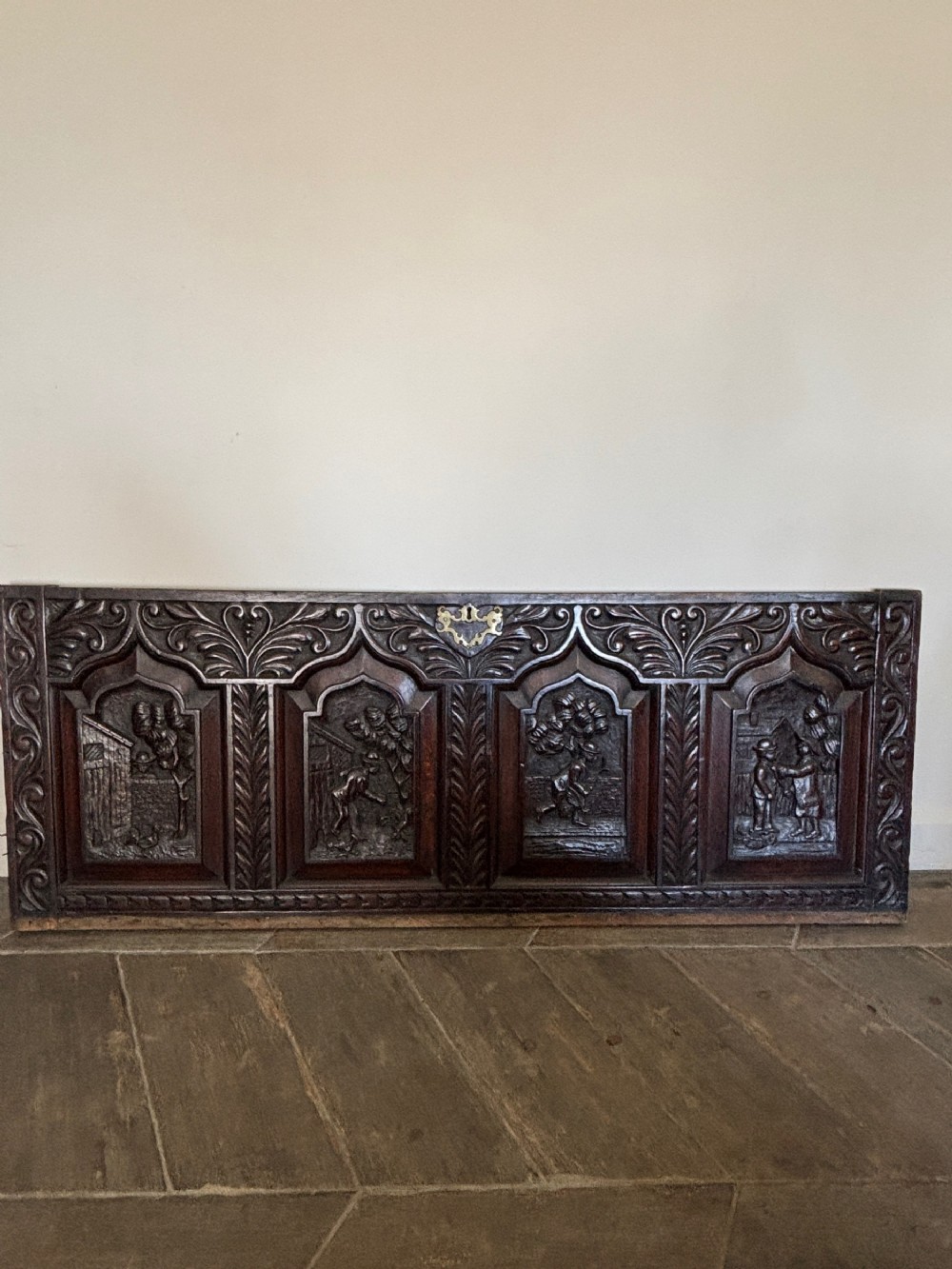 A rectangular dark oak carved panel featuring four pointed arch sections depicting figurative relief scenes with a central brass escutcheon.