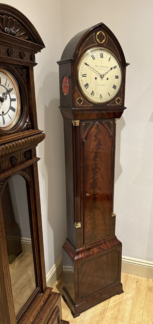 fig 1. A Regency flame mahogany longcase clock featuring a pointed arch hood, white circular dial signed by Thomas Wightman of London, and brass column details.