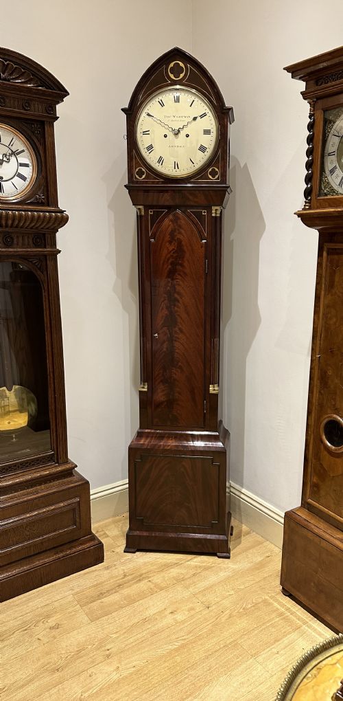 fig 2. A Regency flame mahogany longcase clock featuring a pointed arch hood, white circular dial signed by Thomas Wightman of London, and brass column details.