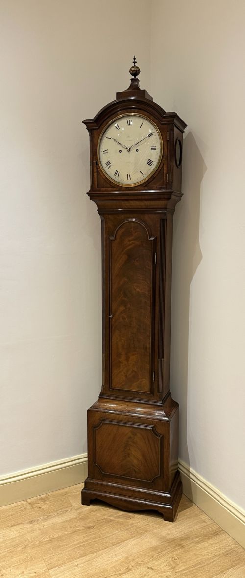 fig 1. A tall Georgian flame mahogany longcase clock featuring an arched hood, brass finial, and a circular white dial with Roman numerals.