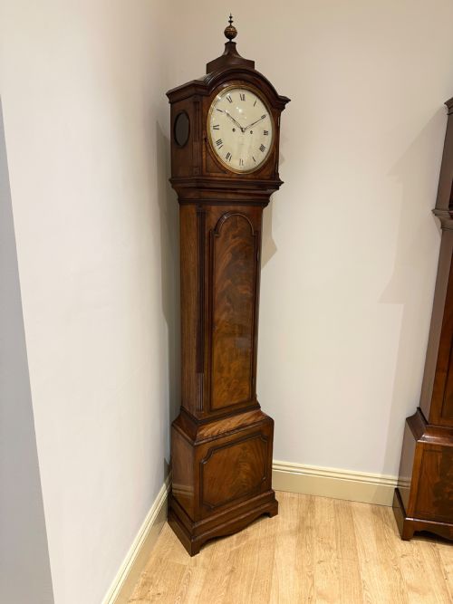 fig 2. A tall Georgian flame mahogany longcase clock featuring an arched hood, brass finial, and a circular white dial with Roman numerals.
