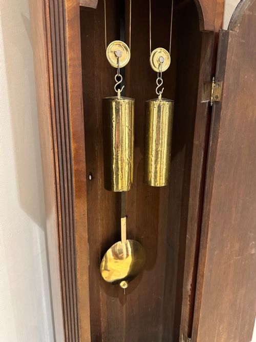 fig 11. A tall Georgian flame mahogany longcase clock featuring an arched hood, brass finial, and a circular white dial with Roman numerals.