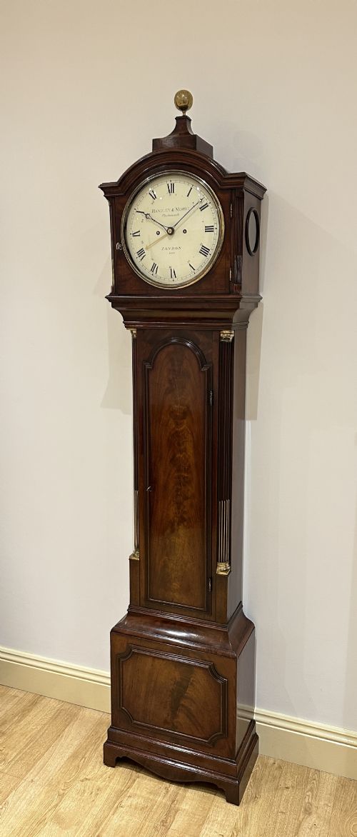 fig 2. A tall Georgian flame mahogany longcase clock featuring an arched dial hood topped with a brass ball finial, reeded side columns, and a shaped trunk door.