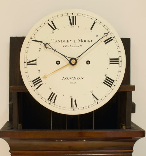fig 7. A tall Georgian flame mahogany longcase clock featuring an arched dial hood topped with a brass ball finial, reeded side columns, and a shaped trunk door.