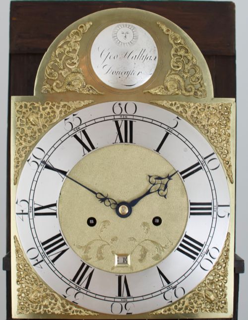 fig 1. An 18th-century Georgian longcase clock featuring a dark wooden cabinet, an arched hood with a finial, and a brass dial with silvered chapter ring.