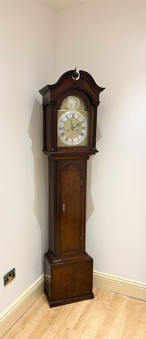 fig 2. An 18th-century Georgian longcase clock featuring a dark wooden cabinet, an arched hood with a finial, and a brass dial with silvered chapter ring.