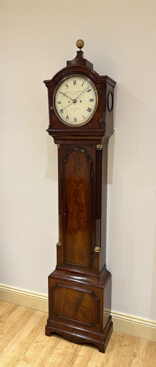 fig 2. A Georgian flame mahogany longcase clock featuring an arched bonnet top with a brass ball finial, reeded columns, and an enamelled circular dial signed Handley and Moore Clerkenwell London.