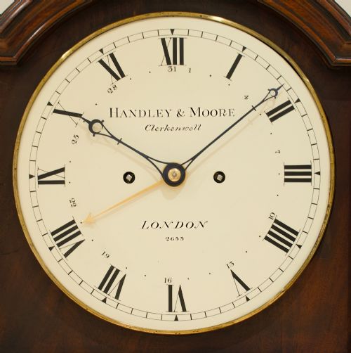 fig 6. A Georgian flame mahogany longcase clock featuring an arched bonnet top with a brass ball finial, reeded columns, and an enamelled circular dial signed Handley and Moore Clerkenwell London.