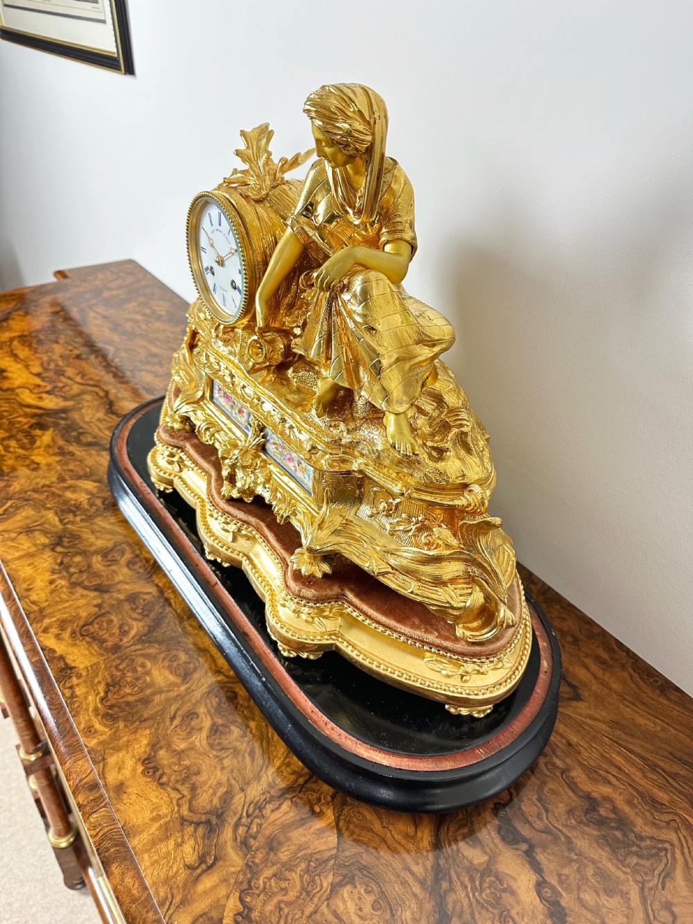 A nineteenth-century French gilt bronze mantel clock featuring a draped classical female figure beside a circular white dial with floral porcelain panels. View X