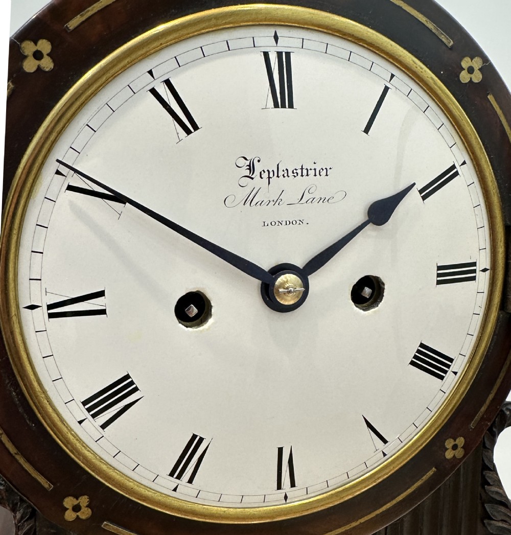 A Regency mahogany and brass bracket clock featuring a round white dial with Roman numerals, topped with a brass pineapple finial and resting on ball feet. View X