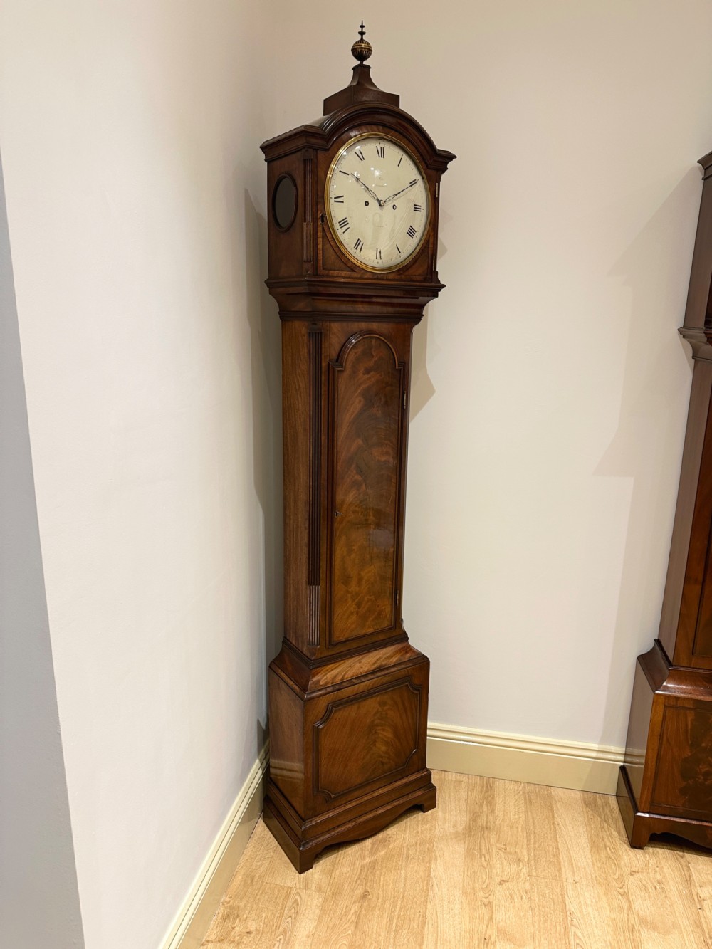 A tall Georgian flame mahogany longcase clock featuring an arched hood, brass finial, and a circular white dial with Roman numerals. View III