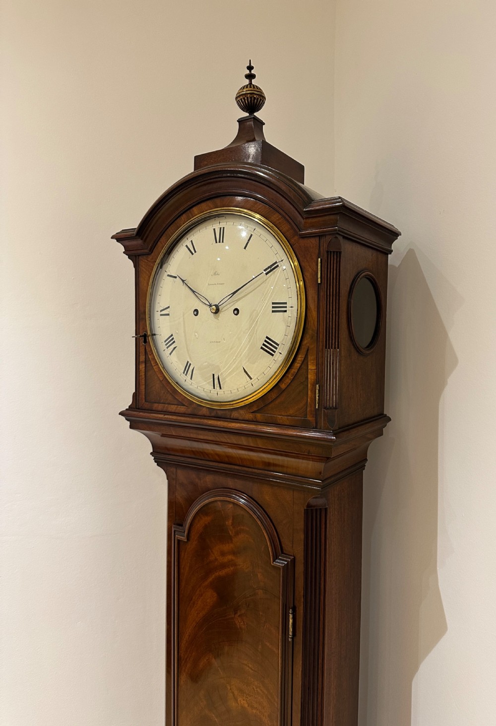 A tall Georgian flame mahogany longcase clock featuring an arched hood, brass finial, and a circular white dial with Roman numerals. View XV