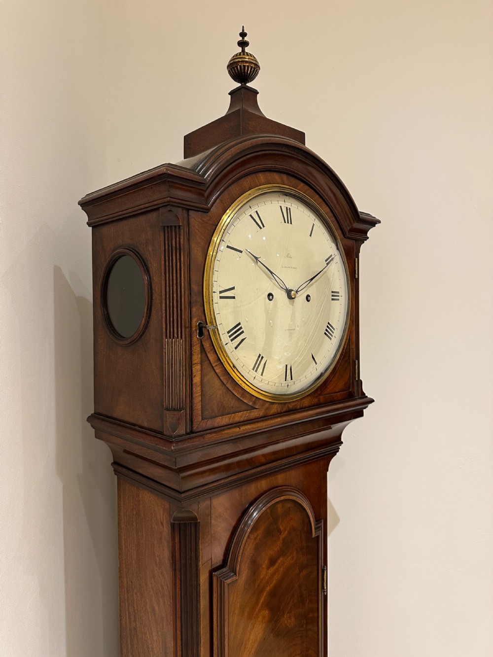 A tall Georgian flame mahogany longcase clock featuring an arched hood, brass finial, and a circular white dial with Roman numerals. View XVI