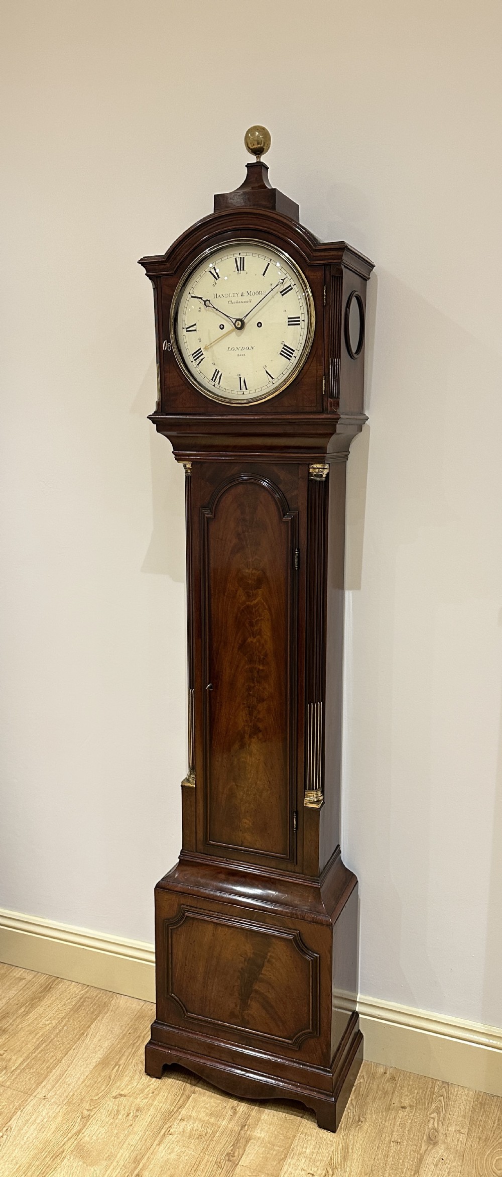 A tall Georgian flame mahogany longcase clock featuring an arched dial hood topped with a brass ball finial, reeded side columns, and a shaped trunk door. View III