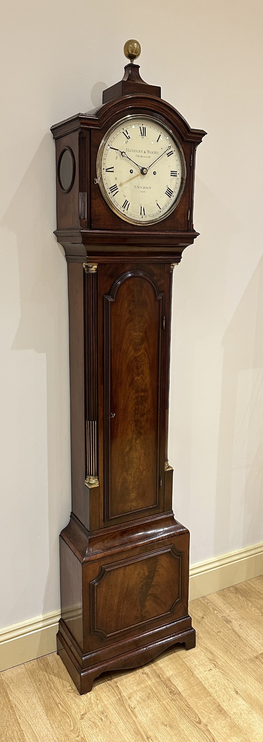 A tall Georgian flame mahogany longcase clock featuring an arched dial hood topped with a brass ball finial, reeded side columns, and a shaped trunk door. View IV