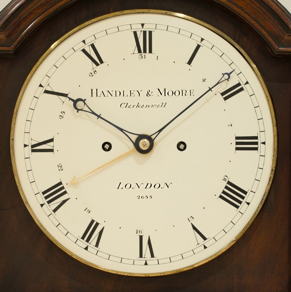 A tall Georgian flame mahogany longcase clock featuring an arched dial hood topped with a brass ball finial, reeded side columns, and a shaped trunk door. View VI