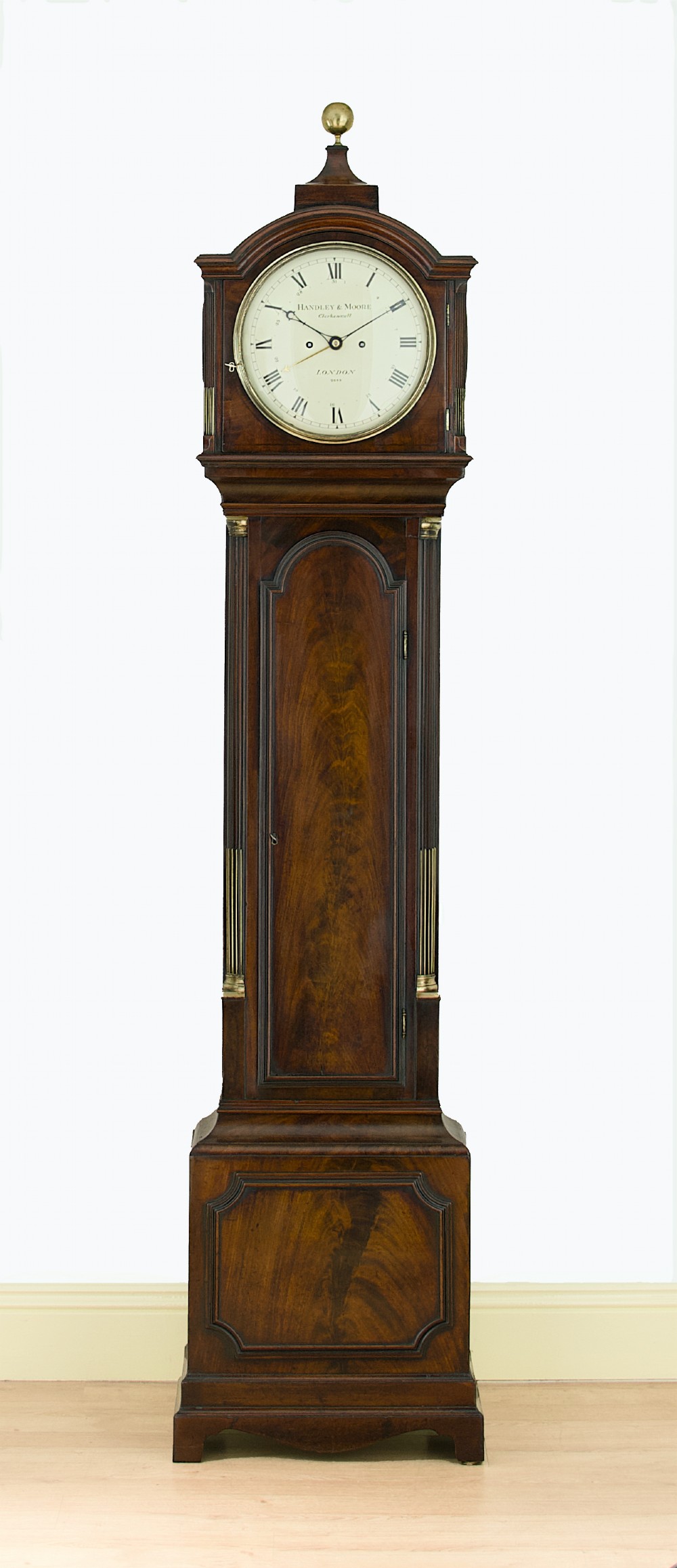 A tall Georgian flame mahogany longcase clock featuring an arched dial hood topped with a brass ball finial, reeded side columns, and a shaped trunk door.