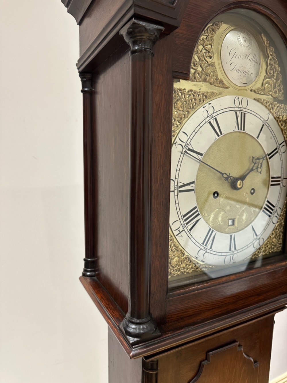 An 18th-century Georgian longcase clock featuring a dark wooden cabinet, an arched hood with a finial, and a brass dial with silvered chapter ring. View VII